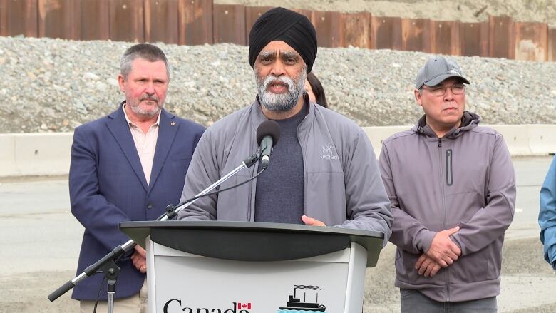 A man stands speaking at a podium outdoors, with two other people looking on.