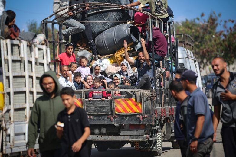 An open-bed truck containing about a dozen people and scores of items and belongings is shown on a roadway as other people walk near the vehicle.