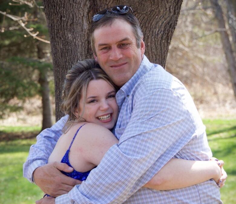 An older man hugs a younger woman wearing a formal blue dress. He has sunglasses on the top of his head. 