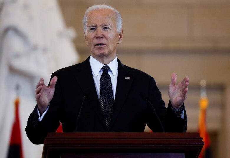 An older cleanshaven man in a suit and tie holds his hands apart while speaking at a podium.