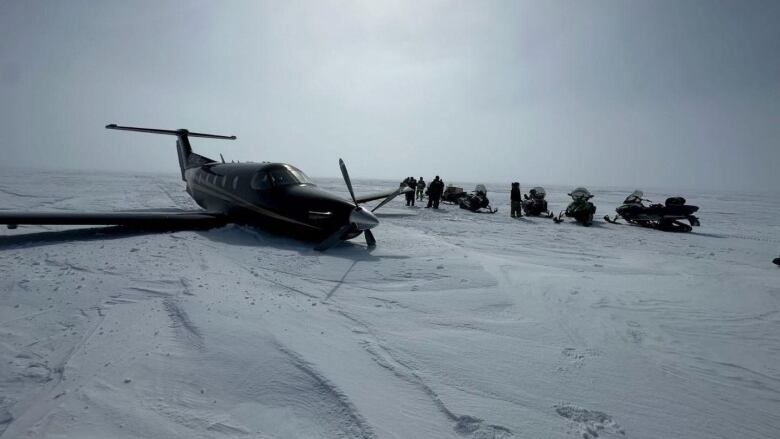 A black plane with a propeller on the front lies upright in the snow and several snowmobiles and people are seen in the background. 