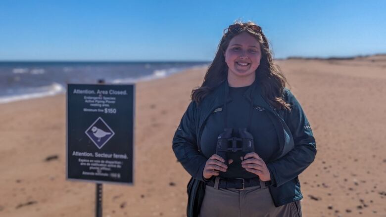 A woman wearing a green Parks Canada uniform and binoculars stands on a beach. 