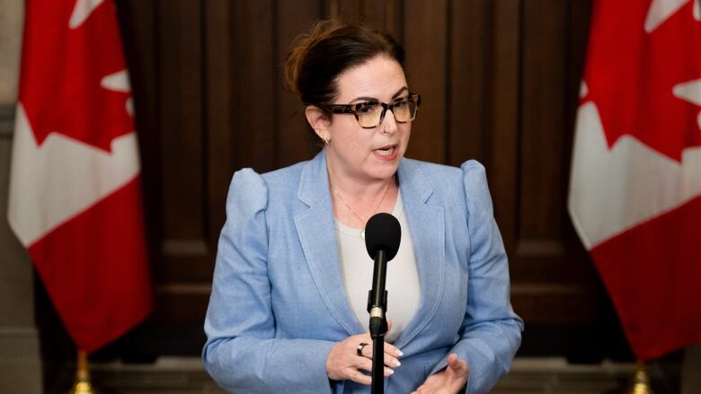 A woman in a blue blazer speaks into a microhpone. Two Canadian flags are draped in the background.