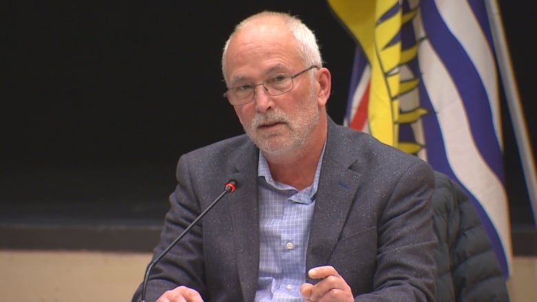 A man with short white hair and a short white beard and glasses speaks into a microphone.