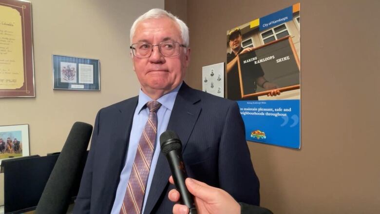 A white man wearing a suit speaks to reports, with a City of Kamloops poster behind him.