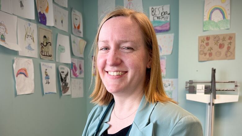 A woman with blond hair wearing a pale green jacket and black shirt with a stethoscope around her neck stands in front of a wall covered with children's drawings.