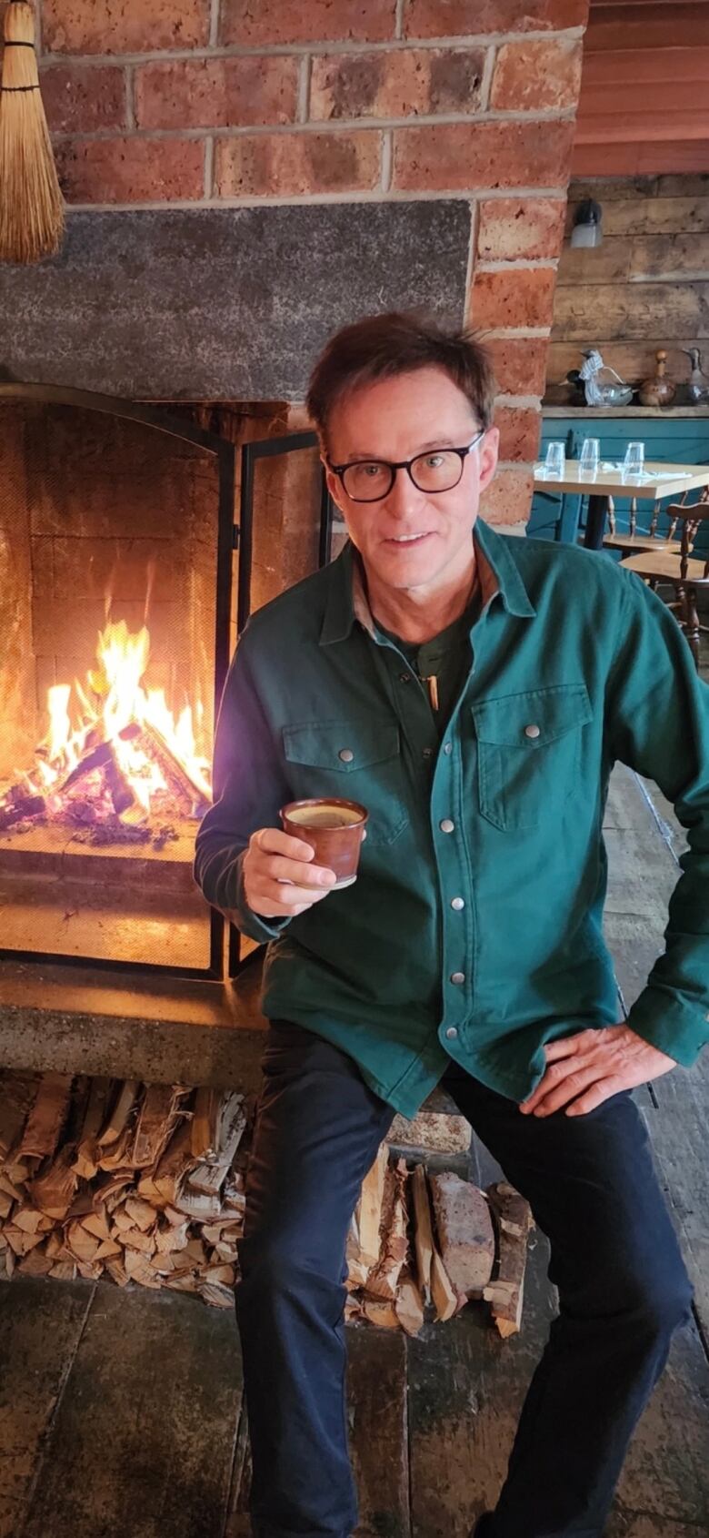 Man with glasses sitting in front of a brick fireplace holding glass.