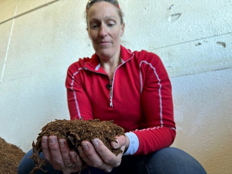 A woman with a red long sleeve shirt holds up a handful of brown material. It looks like dirt and small wood chips.