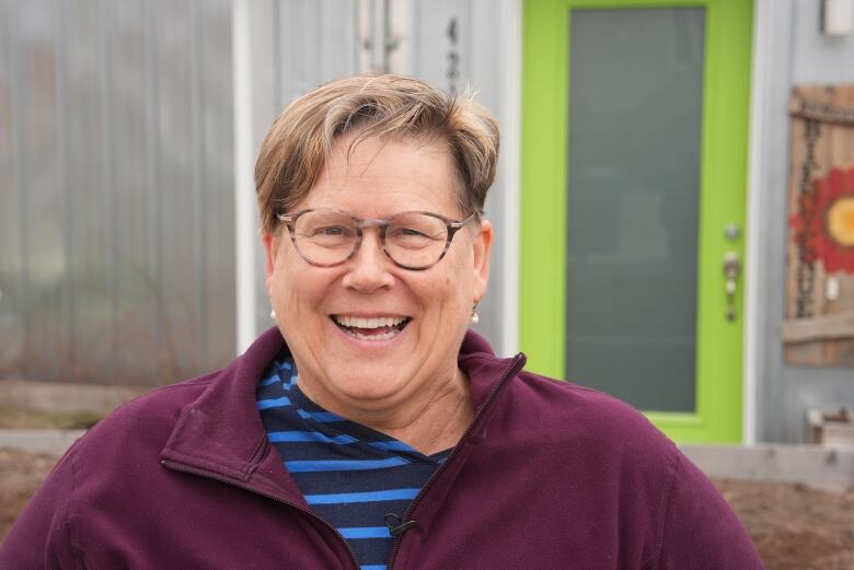 A woman with short hair and glasses laughing. She's outside, in front of a building with a green door.