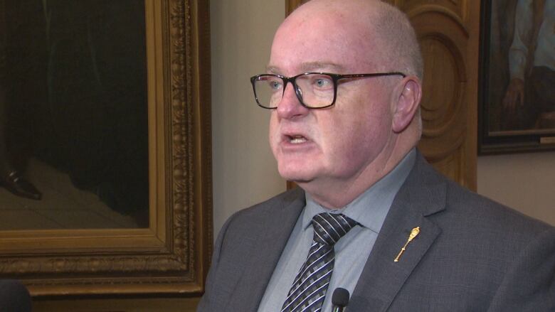 A man wearing glasses, a blazer, collared shirt and tie, speaking.