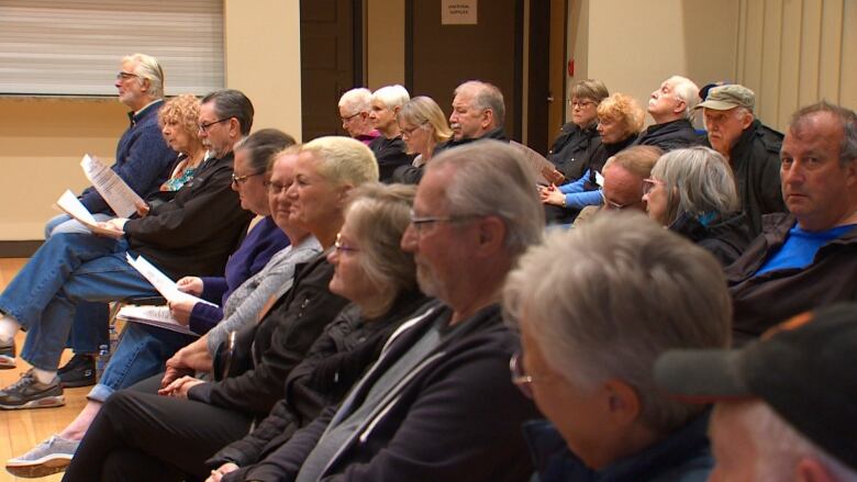 A side-view of a group of people seated. They are listening to and watching a meeting.