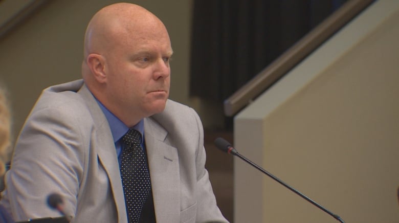 A bald man in a suit looks forward from a desk at a council meeting.