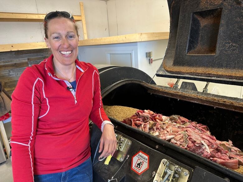 A woman in a red long sleeve grins at the camera. She's leaning against a big black cylinder with an open hatch, inside you can see a pile of fish guts on top of brown grain.