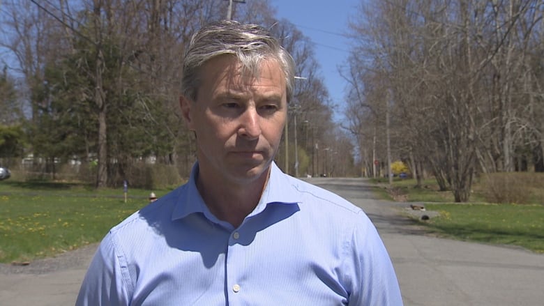 Man wears a blue button-up shirt stands in a park on a sunny day.