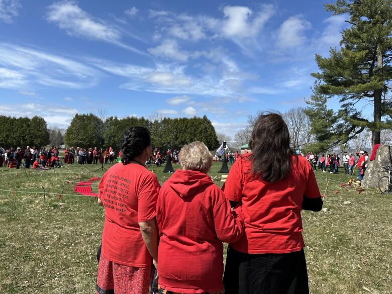 Three women dressed in red clothing stand next to each other outside on a grassy field with their backs to the camera.