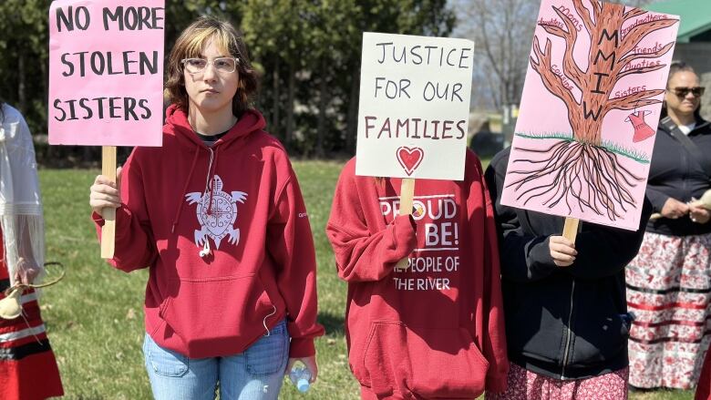 A few people wearing red stand outside in the sun holding up hand-made signs.