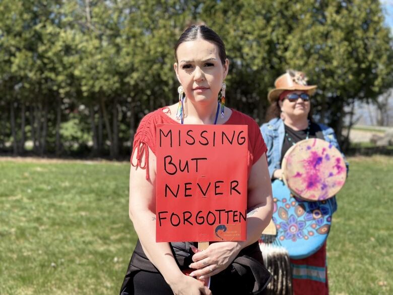 A young woman with slicked back, dark hair stands outside holding a sign that says missing but never forgotten.