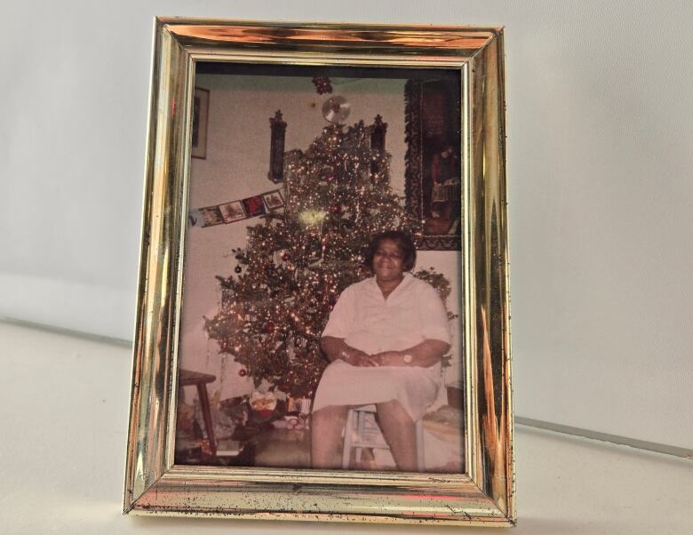 A gold photo frame that holds an old photo of a woman sitting in front of a Christmas tree.