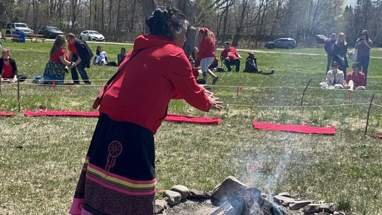 A woman wearing a red sweater and long skirt stands over a fire, rubbing her hands in the smoke.