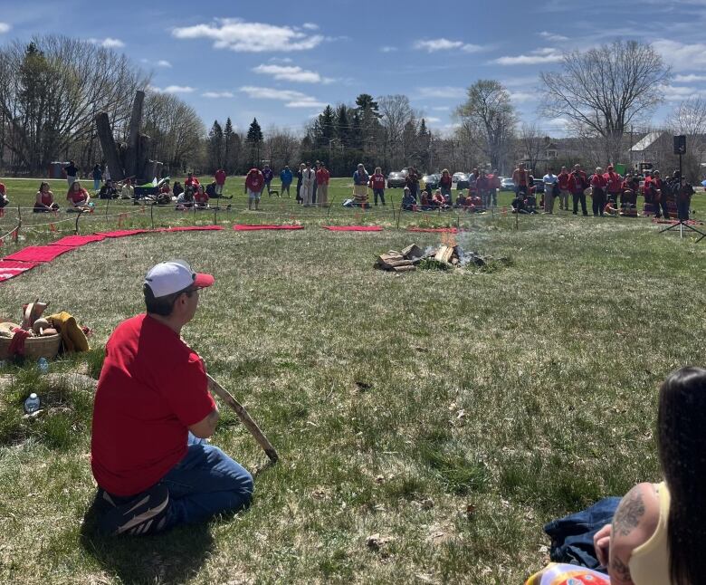 A group of people sit in a wide circle around a smoking fire on a sunny day.