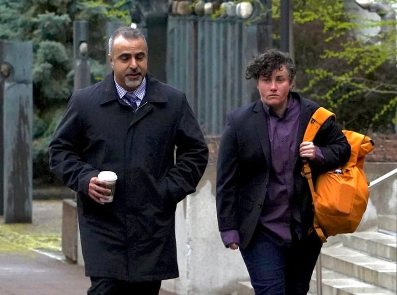 Two people  one carrying an orange bag, the other holding a coffee cup  walk down a city street on a cloudy day.