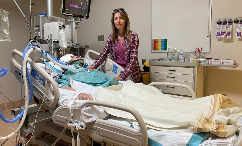 Woman lying in hospital bed. Woman with blond hair wearing purple stands beside her