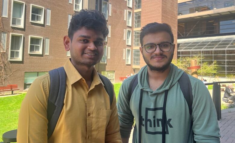 Two young men pose for the camera in front of a grassy area with university buildings in the background.