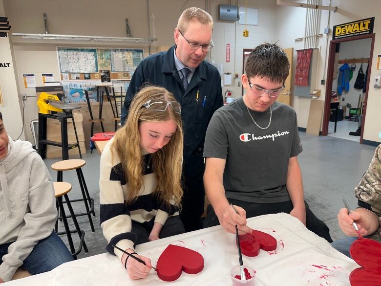 A teacher is shown in a shop class in the middle of two students who are painting wooden hearts red.