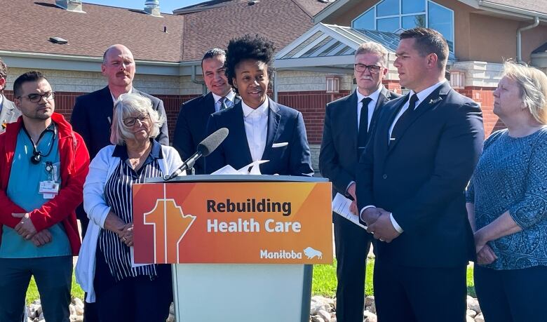 A group of ten politicians and their supporters stand behind a lectern and smile at an outdoors government news announcement.