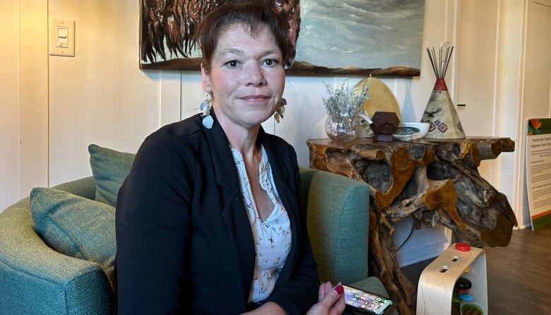 Shannon Johansen is seen sitting on a chair in a room with a buffalo painting with indigenous displays behind her.