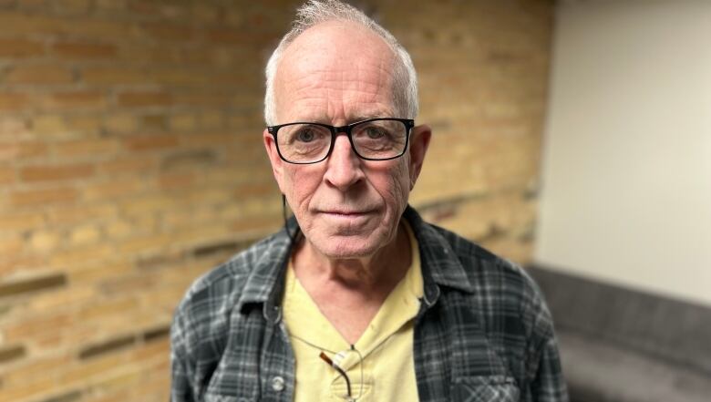 An elderly man wearing glasses and a yellow polo shirt stands in a room with an exposed brick wall. He's looking right at the camera with a neutral face. 