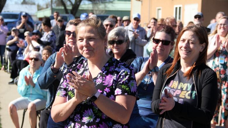 A group of dozens of people clap their hands outside in a parking lot in the summer.