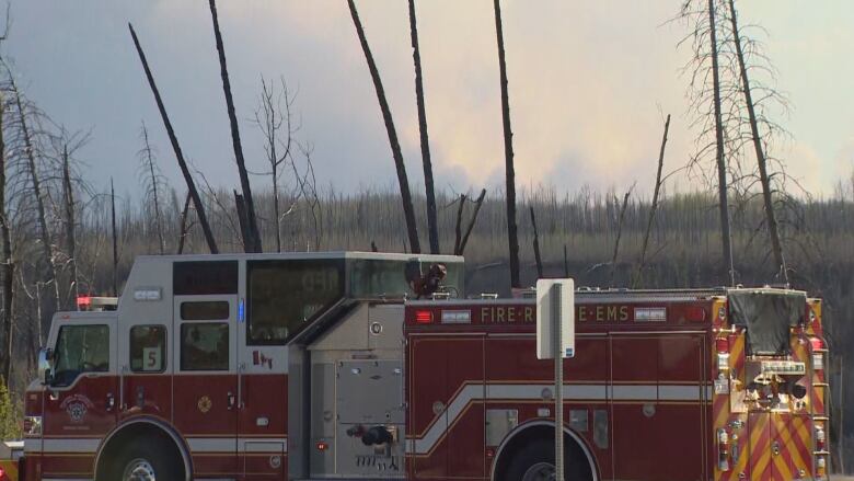 A fire truck sits in the foreground, with a wooded area behind it. Large smoke plumes float in the air in the background.