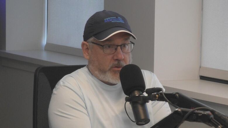 A person wearing a white shirt and black baseball cap speaking into a microphone.