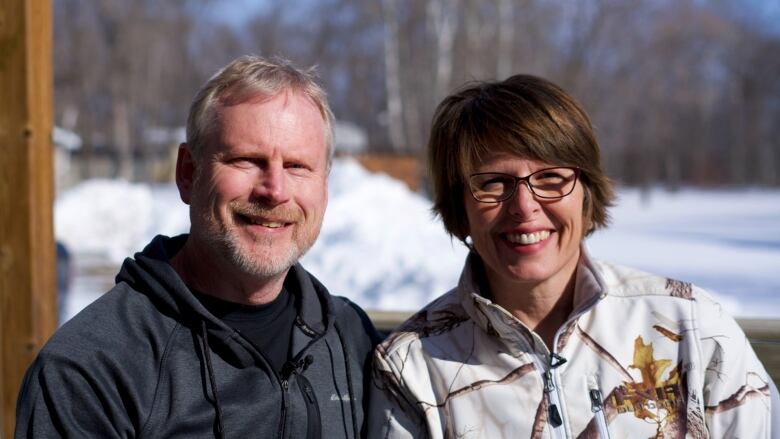 A man with blond hair and a blond and grey beard smiles while sitting next to a smiling woman with brown ear-length hair and black-framed glasses.