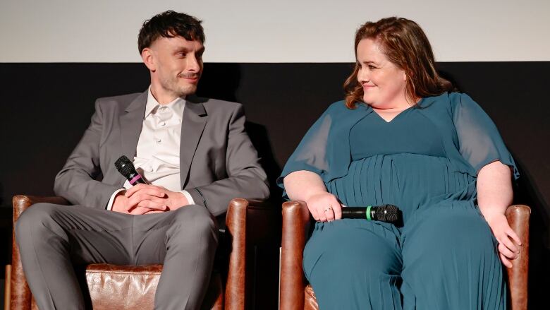 A man in a suit and a woman in  a green dress smile at each other while sitting on a stage