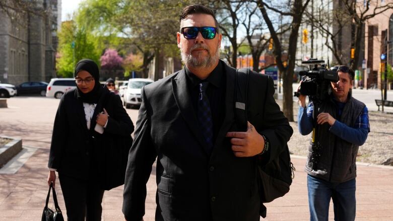 A man walks outside in a suit and sunglasses.