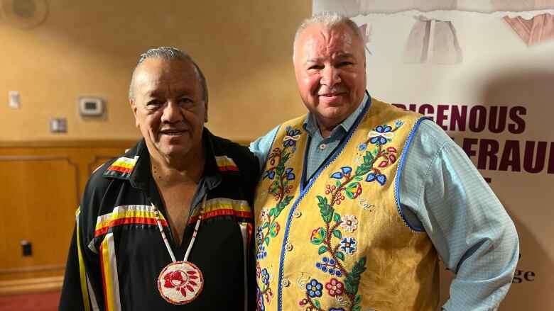 Two men, one in a First Nations ribbon shirt the other in a Mtis beaded vest, pose for a picture.