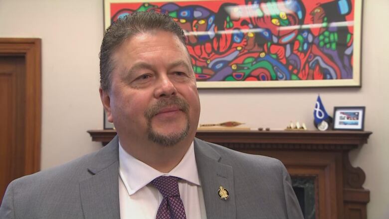 A man with short grey hair and a goatee wearing a grey blazer and purple tie stands in an office.