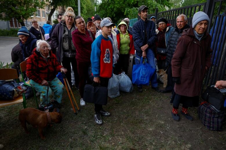 Several people of all ages are shown standing outdoors, most wearing jackets and coats while carrying bags and belongings.