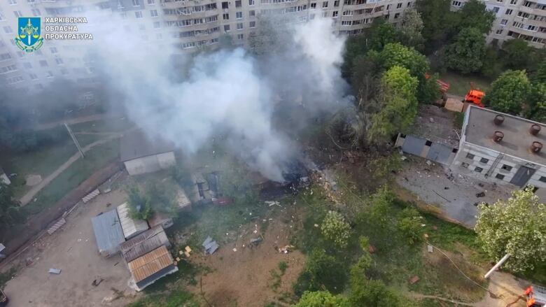 An overhead shot shows the ground between hirise buildings, with smoke seen rising from a leafy area.