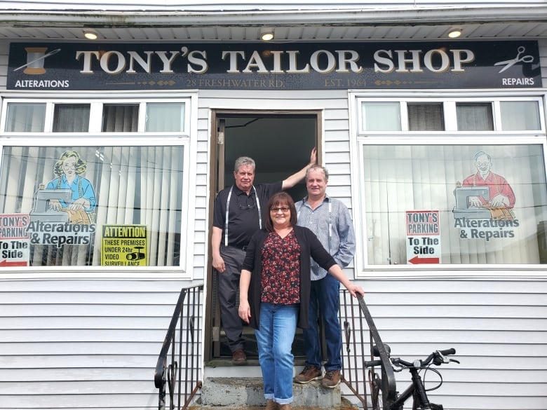 Two men and a woman stand on the front steps of a white building. Above them, in white letters on a dark background, a sign reads 'Tony's Tailor Shop.'