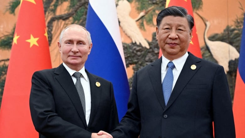 Two men in business suits and ties grip hands and look at the camera in front of flags.