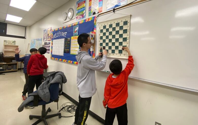 Two boys plot pawns on a chess board hung on a wall.