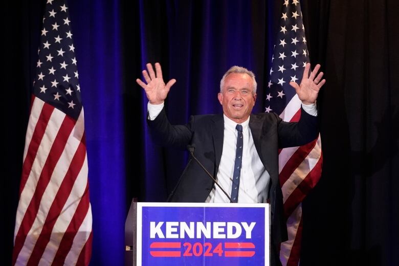 A man wearing a black suit jacket, white shirt and black tie waves his hands while standing in front of two red, white and blue U.S. flags and behind a podium with a sign reading 