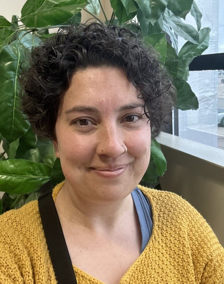 A smiling woman with short, brown hair and wearing a yellow sweater sits in front of a leafy green plant.