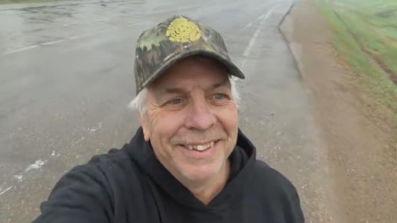 A man in a trucker hat stand on a road smiling.