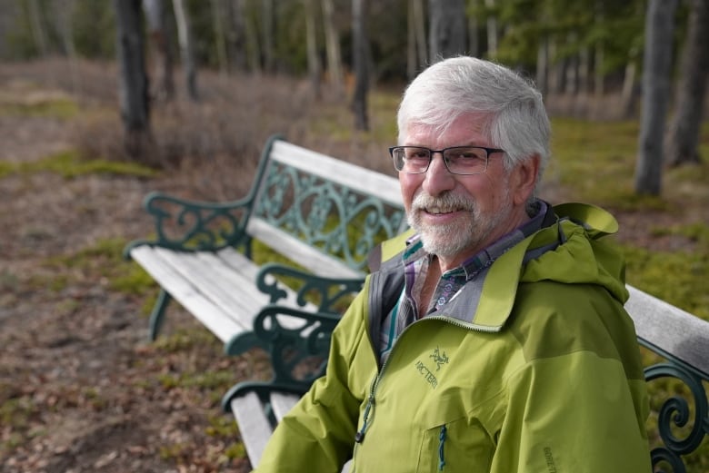 A man with white hair and beard sits on a bench in the forest.