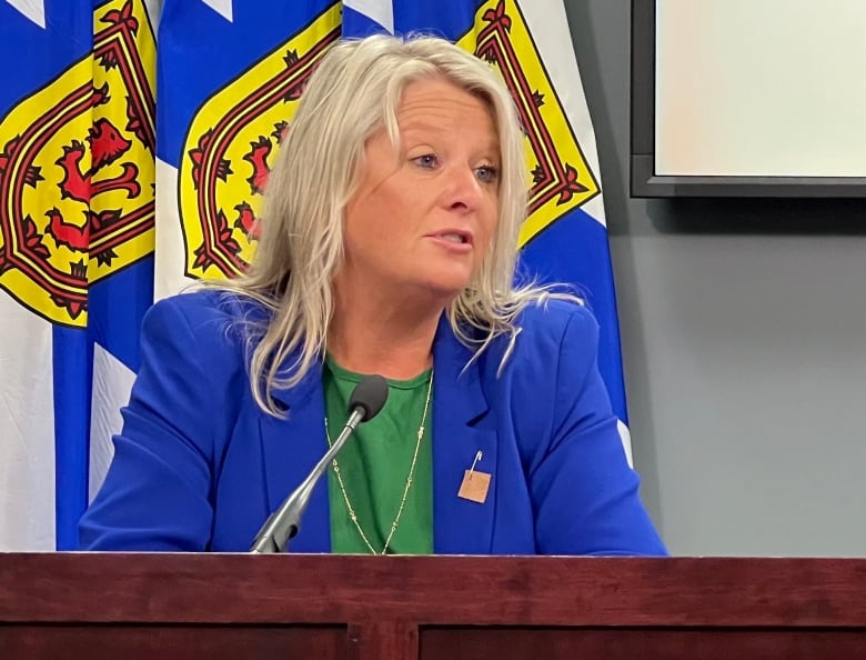 A woman wearing a blue blazer sits at a desk in front of a microphone. There are Nova Scotia flags behind her.