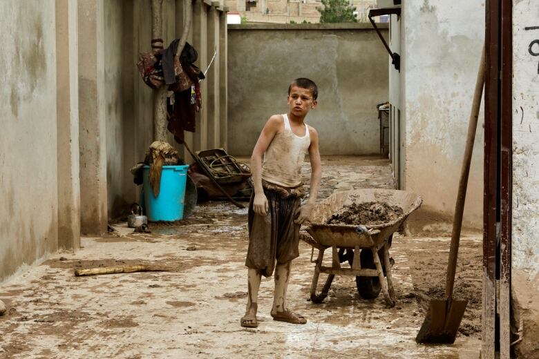 A boy wearing shorts and a tank top caked in mud stands in an alley holding a wheelbarrow full of mud. 